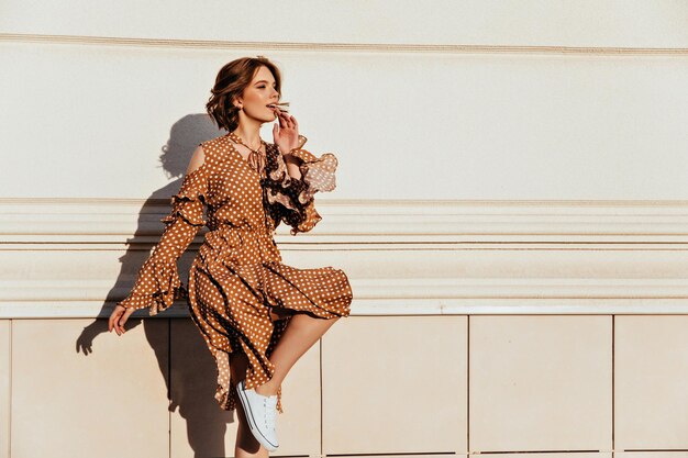 Geweldig wit meisje met golvend haar dat danst in de buurt van de muur Buitenopname van een mooi elegant vrouwelijk model draagt een bruine jurk