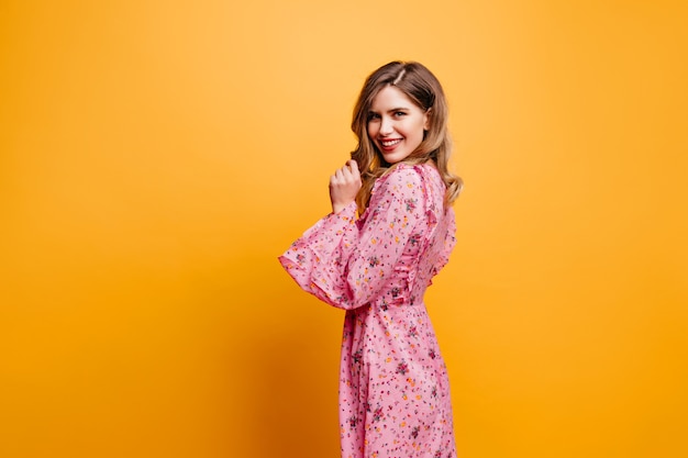 Geweldige blanke vrouw in lange jurk kijkt over de schouder. Jocund krullend meisje in roze outfit poseren op gele muur.