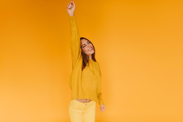 Geweldige brunette dame in stijlvolle gele broek die geluk uitdrukt blij slank meisje poseren met hand omhoog op oranje achtergrond