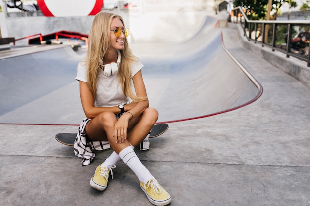 Gratis foto geweldige vrouw in grote koptelefoon zittend op skateboard. openluchtportret van het mooie blonde vrouwelijke model stellen met longboard.