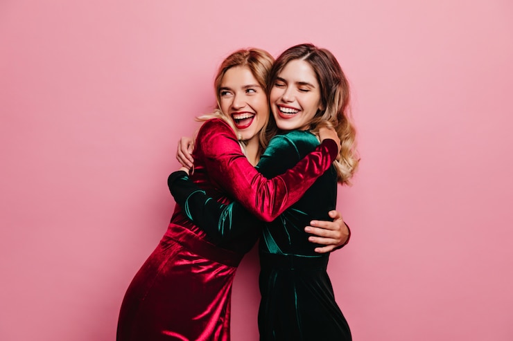 Gratis Foto geweldige vrouw in rood fluwelen jurk omarmen haar zus. verfijnde meisjes die plezier hebben op een roze muur.