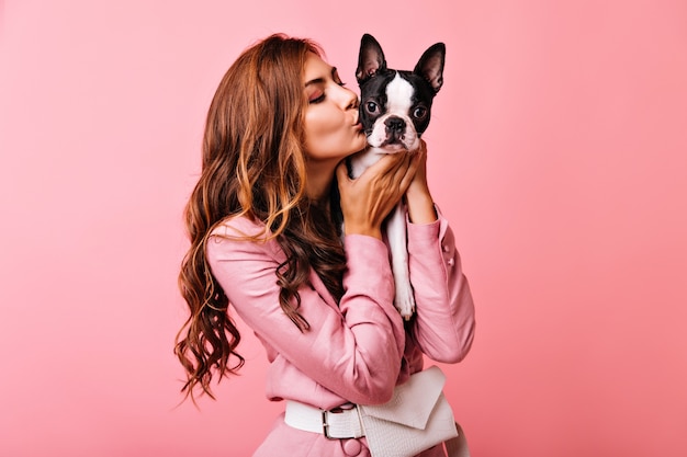 Geweldige vrouw met lang golvend haar Franse bulldog kussen. portret van gember meisje omarmen haar puppy op roze.