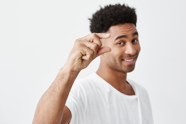 Gewoon een klein beetje. Zachte focus. Geïsoleerde portret van jonge knappe donkere man met krullend haar in casual wit t-shirt gebarend met hand, in de camera kijken met blij en ontspannen gezicht