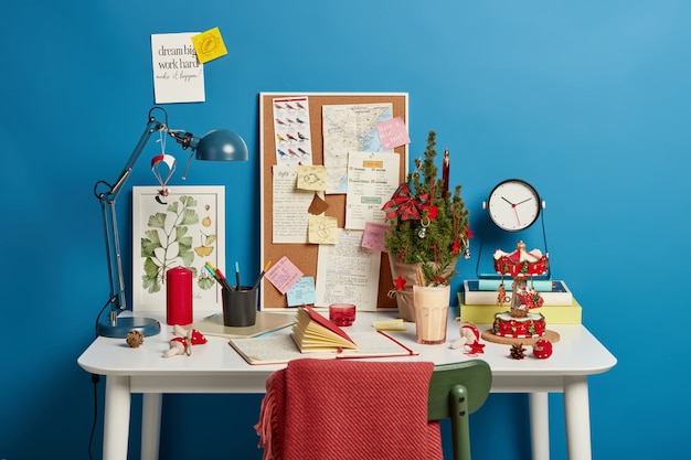 Gezellige coworking-ruimte met blocnote, versierde dennenboom, gekoelde zoete dagelijkse drank, rode onverbrande kaars, stoel met plaid, handgeschreven notities.