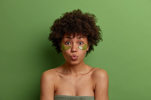 Gezichtsbehandeling concept. Mooie jonge opgefrist vrouw met Afro kapsel, maakt gebruik van groene vlekken onder de ogen, rondt de lippen, staat gewikkeld in een handdoek, heeft natuurlijke schoonheid, vormt over groene muur