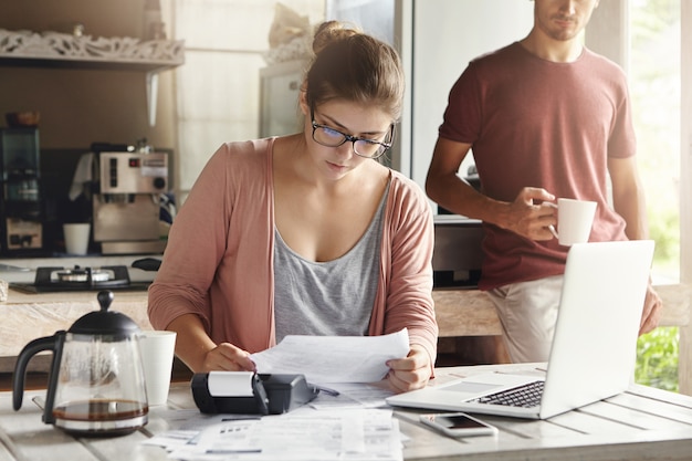 Gezinsbudget en financiën. Jonge vrouw die rekeningen samen met haar echtgenoot thuis doen, die nieuwe aankoop plannen. Ernstig wijfje in glazen die stuk van document houden en noodzakelijke berekeningen maken