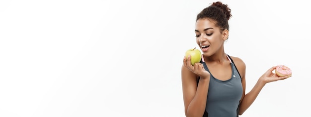 Gratis foto gezond en dieetconcept mooie sportieve afro-amerikaan maakt een beslissing tussen donut en groene appel geïsoleerd op witte achtergrond