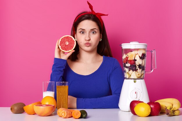 Gezond vegetarisch meisje opent wijd haar ogen, blaast haar mond, kijkt rechtstreeks naar de camera en houdt fruit in één hand. Verschillende fruit is op tafel en in de blender, heerlijke smoothie koken.