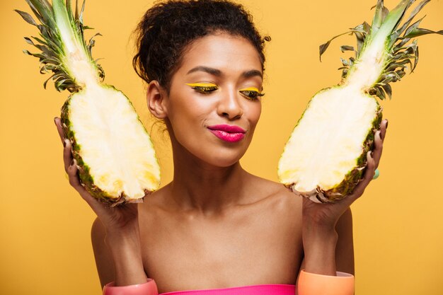 Gezonde gemengd rasvrouw met maniermake-up die detox hebben die verse rijpe ananas houden die in half geïsoleerd, over gele muur wordt verdeeld