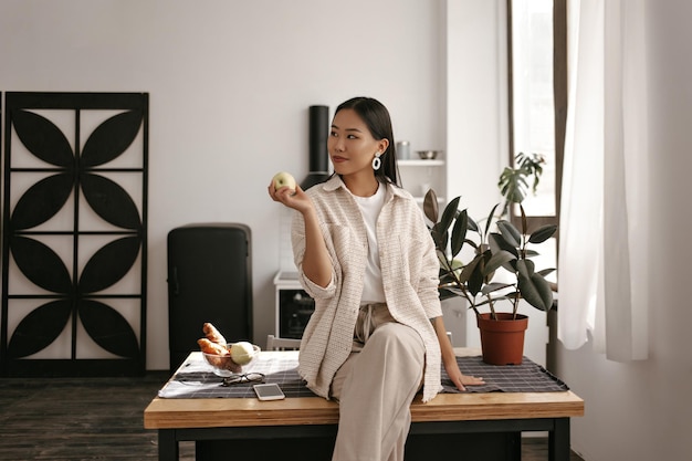 Gezonde jonge brunette Aziatische vrouw in broek en beige vest zit op een houten tafel in de keuken, glimlacht en houdt verse, smakelijke appel