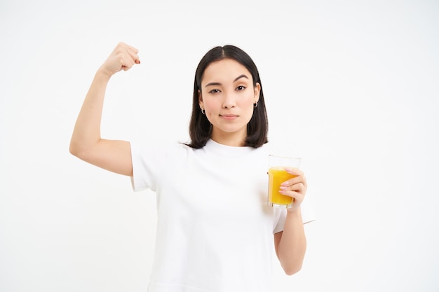 Gratis foto gezonde levensstijl lachende jonge aziatische vrouw toont sterke spieren biceps bij de hand en drinkt jus