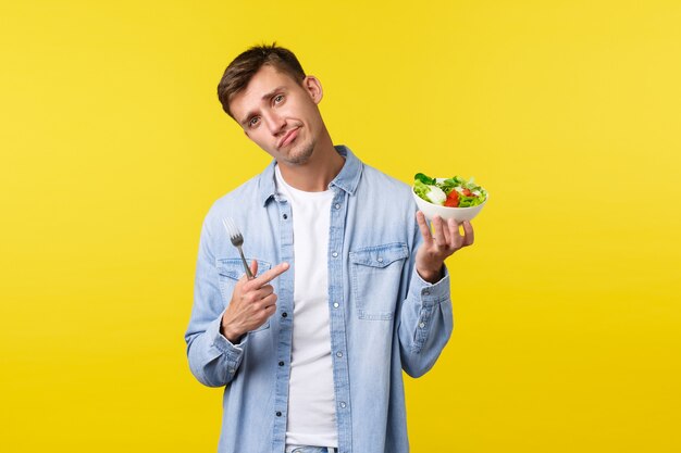 Gezonde levensstijl, mensen en voedselconcept. Aarzelende knappe jongeman die met de vinger wijst naar walgelijke salade, dit niet wil eten, ontevreden grijnzend en hoofd kantelt, droevige, gele achtergrond.