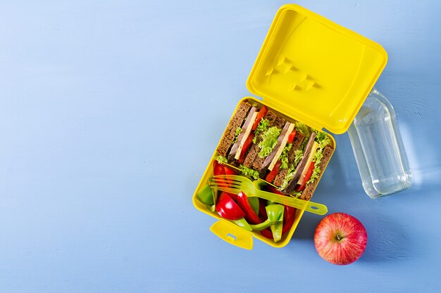 Gezonde school lunchbox met rundvlees sandwich en verse groenten, fles water en fruit op blauwe tafel. Bovenaanzicht. Plat liggen