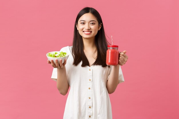 Gezonde voeding, emoties en zomer lifestyle concept. Enthousiast en vrolijk schattig Aziatisch meisje vol energie, smakelijke frisse salade eten en smoothie drinken, glimlachend in de camera gelukkig, roze achtergrond