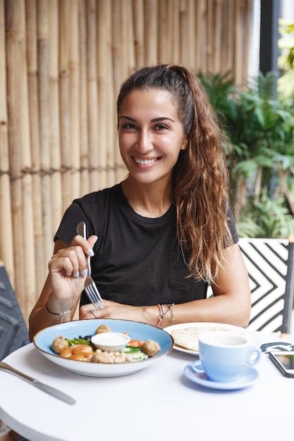 Gezonde vrouw met bruin, zittend in een t-shirt op caféterras, ontbijt eten en koffie drinken.