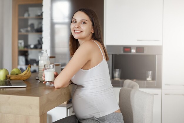 Gezondheids- en moederschapsconcept Aantrekkelijke moeder die wacht op de geboorte van de baby, zittend in de keuken en het drinken van melk die gezond voedsel eet en vrolijk glimlacht naar de camera terwijl ze met haar man over hun zoon praat