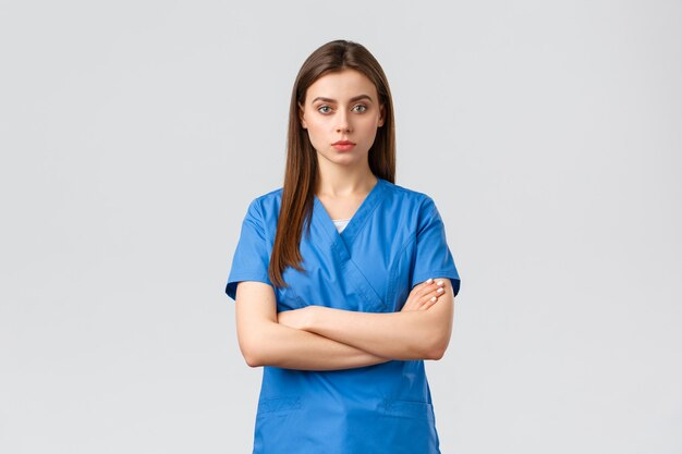 Gezondheidswerkers, voorkom virus-, verzekerings- en medicijnconcept. Ernstige en zelfverzekerde vrouwelijke verpleegster in blauwe scrubs, stagiair of arts kruist de armen op de borst, kijk camera streng, klaar om covid-19 te bestrijden