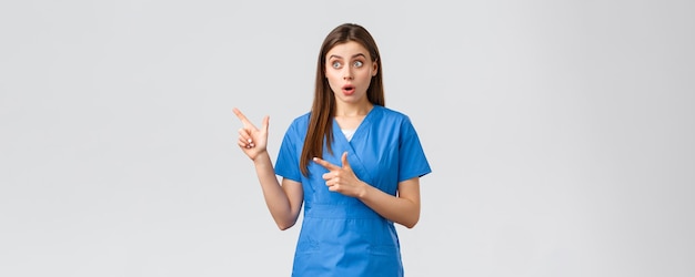 Gezondheidswerkers voorkomen virusverzekering en medicijnconcept Verrast vrouwelijke verpleegster of arts in blauwe scrubs kijken en wijzende vingers linkerbovenhoek hijgend verbaasd