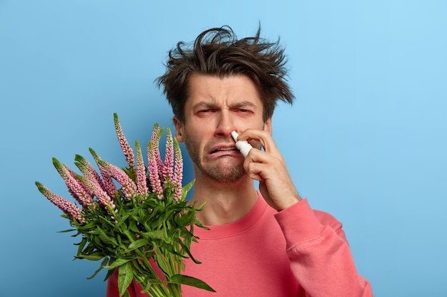 Gezondheidszorg en allergiesymptomen concept. Ontevreden man geneest allergische rhinitis met neusdruppels, heeft een zieke uitdrukking veroorzaakt door trigger, geïrriteerde rode ogen, poseert binnenshuis, is overgevoelig