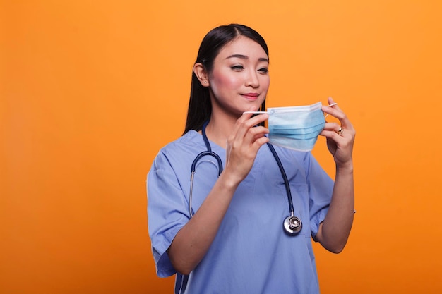 Gezondheidszorgkliniek aziatische verpleegster in blauw uniform, met een stethoscoop die een gezichtsmasker opzet tegen covid. Ziekenhuisverzorger die een virusbeschermend masker opzet terwijl hij een medisch instrument draagt.