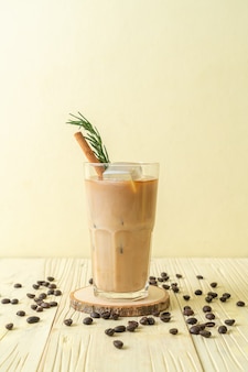 Gietende melk in zwart koffieglas met ijsblokje, kaneel en rozemarijn op houten ondergrond