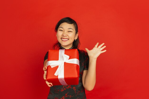 Giftbox geven. Gelukkig Chinees Nieuwjaar 2020. Het portret van het Aziatische jonge meisje dat op rode achtergrond wordt geïsoleerd. Vrouwelijk model in traditionele kleding ziet er gelukkig uit. Viering, vakantie, emoties. Copyspace.