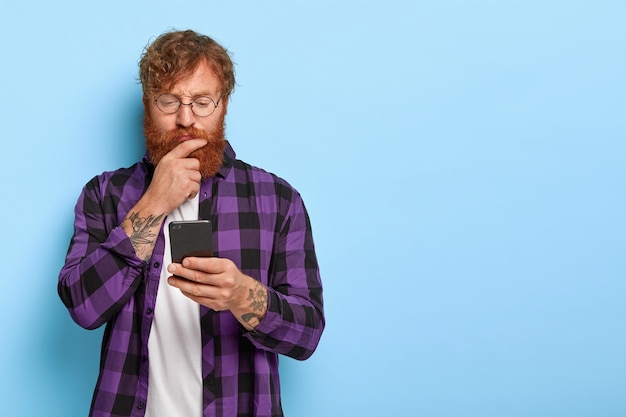 Ginger millennial guy gebruikt mobiele telefoon applicatie voor het synchroniseren van gegevens, heeft een serieuze blik, draagt een ronde bril