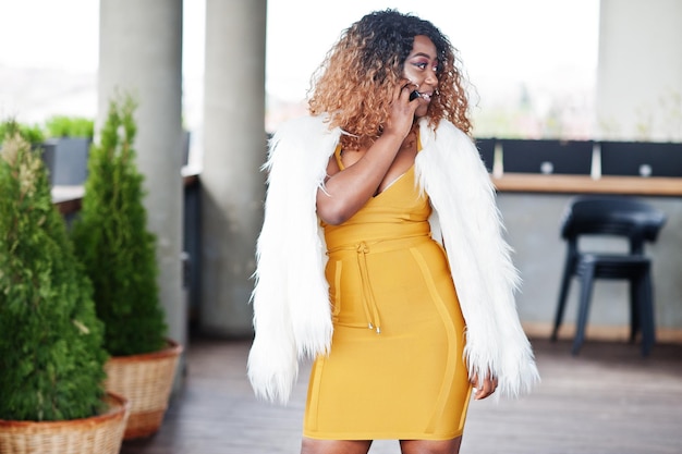 Glamour afro-amerikaanse vrouw in gele jurk en witte wollen cape poseerde op balkon en praat op mobiele telefoon