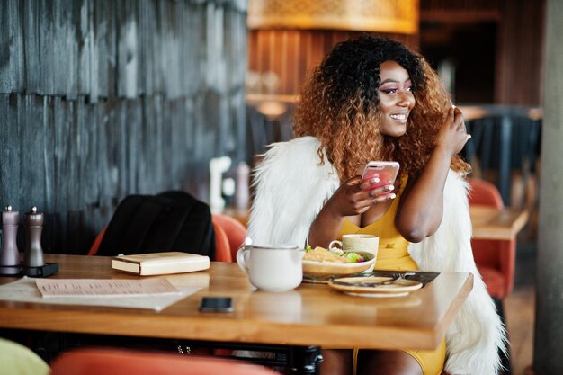 Glamour Afro-Amerikaanse vrouw in gele jurk zittend aan tafel met gerechten op restaurant met mobiele telefoon bij de hand