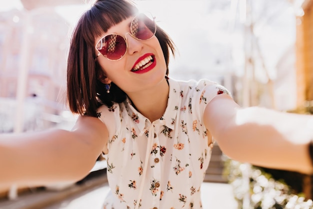 Glamoureuze vrouw in zomerbril die selfie maakt in zonnige ochtend Buitenopname van zalige brunette meisje draagt witte blouse