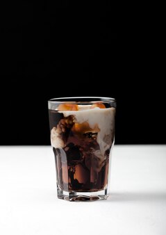 Glas zwarte koffie met ijsblokjes en verse melk op wit bord en zwarte achtergrond.