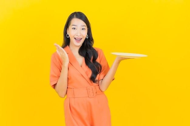 Glimlach van de portret de mooie jonge Aziatische vrouw met lege plaatschotel op geel