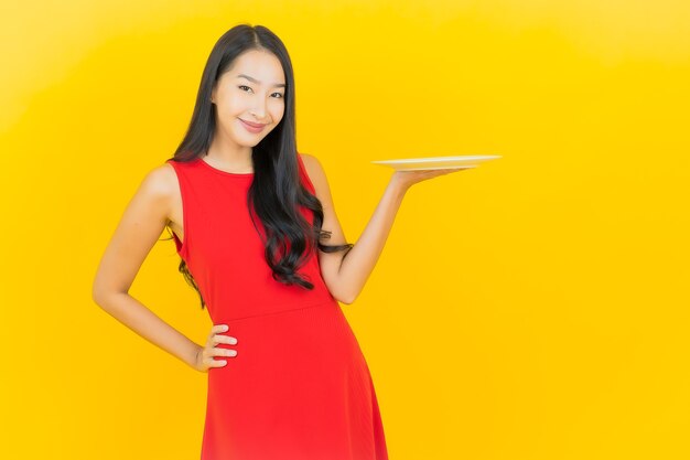 Glimlach van de portret de mooie jonge aziatische vrouw met lege plaatschotel op gele muur