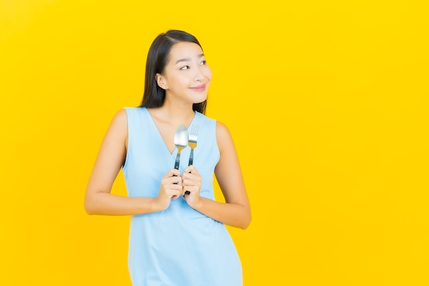 Glimlach van de portret de mooie jonge Aziatische vrouw met lepel en vork op gele kleurenmuur