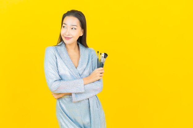 Glimlach van de portret maakt de mooie jonge aziatische vrouw met schoonheidsmiddel omhoog borstel op gele muur