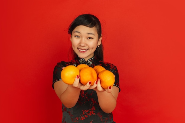 Glimlachen, mandarijnen geven. gelukkig chinees nieuwjaar. aziatisch jong meisje portret op rode achtergrond. vrouwelijk model in traditionele kleding ziet er gelukkig uit. copyspace.