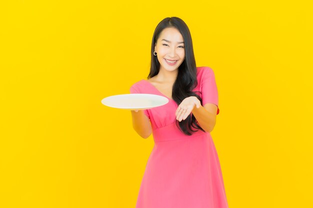 Glimlachen van de portret de mooie jonge Aziatische vrouw met lege plaatschotel op gele muur