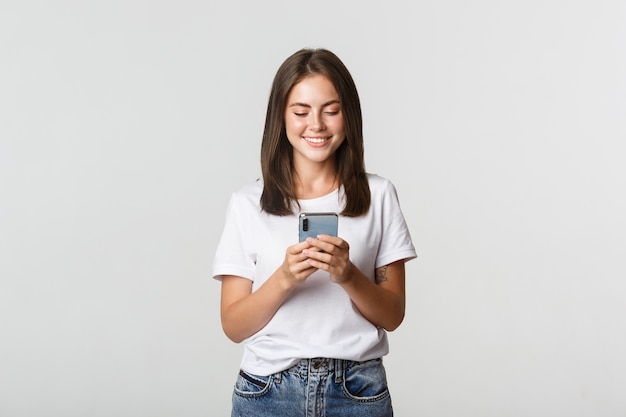 Glimlachend aantrekkelijk meisje dat mobiele telefoon met behulp van en tevreden scherm bekijkt.