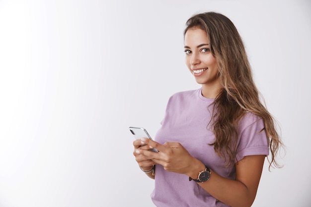 Glimlachend aantrekkelijke gekrulde gebruinde kaukasische 25s vrouw met smartphone half gedraaid draaiende camera glimlachend op mysterieuze wijze wil hulp vragen met behulp van gloednieuwe gadget app witte achtergrond