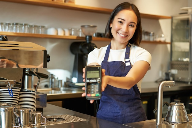 Gratis foto glimlachend aziatisch barista-koffieshoppersoneel geeft u creditcardverwerkingsmachine met pos te