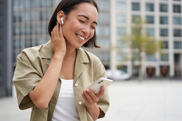 Glimlachend aziatisch meisje luistert naar muziek in draadloze hoofdtelefoons kijkt naar haar telefoon en kiest muziek of podcast