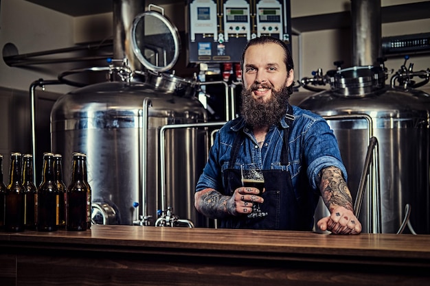 Glimlachend bebaarde getatoeëerde hipster man in een jeans shirt en schort werken in een brouwerij fabriek, staande achter een toonbank, houdt een glas bier vast voor kwaliteitscontrole.