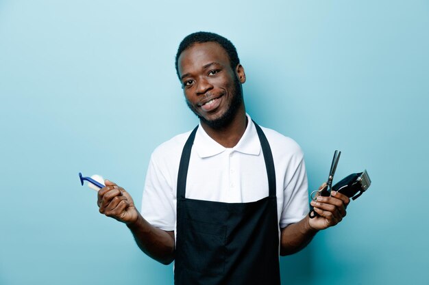 Glimlachend bedrijf kapper tools jonge Afro-Amerikaanse kapper in uniform geïsoleerd op blauwe achtergrond