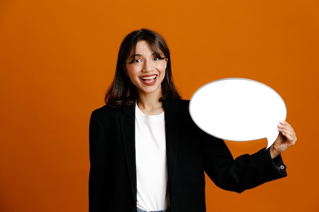 Glimlachend bedrijf tekstballon jonge mooie vrouw draagt zwarte jas geïsoleerd op een oranje achtergrond