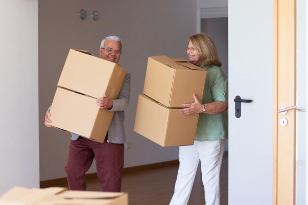 Glimlachend bejaarde echtpaar verhuizen naar nieuw huis. Gepensioneerde man en vrouw houden kartonnen dozen vast en dragen dingen. Onroerend goed, aankoop, verhuisconcept