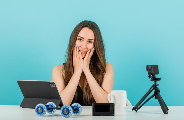 Glimlachend bloggermeisje kijkt naar de camera door handen op de wangen te houden op een blauwe achtergrond