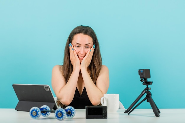 Glimlachend bloggermeisje kijkt naar haar kleine camera door handen op wangen op blauwe achtergrond te leggen