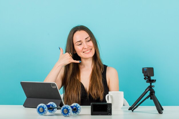 Glimlachend bloggermeisje poseert door telefoongebaar met hand op blauwe achtergrond te tonen