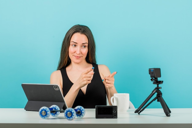 Gratis foto glimlachend bloggermeisje poseert voor de camera door de camera met wijsvingers op een blauwe achtergrond te richten