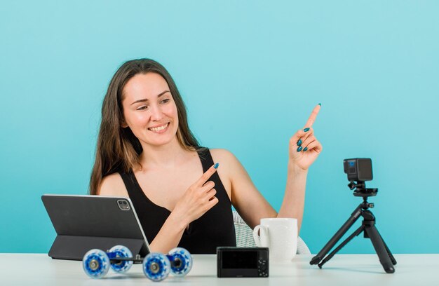 Glimlachend bloggermeisje poseert voor haar kleine camera door met wijsvingers naar rechts te wijzen op een blauwe achtergrond